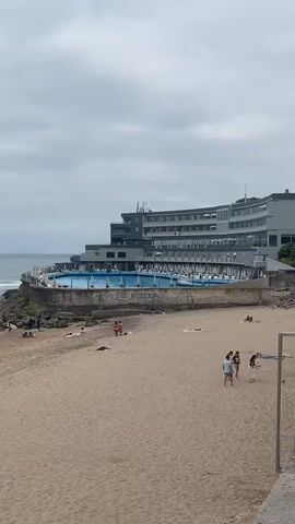 The media file displays a panoramic view of a beach and a hotel complex overlooking the ocean. The beach is sandy, with several people scattered across it, some sunbathing, others walking or playing. There are beach chairs and umbrellas for shade.

Dominating the upper portion of the frame is a modern hotel with multiple levels and a curved facade. A large swimming pool with blue water and loungers is situated on a terrace between the hotel and the beach, offering a view of the sea. Waves are breaking on the shore, and a few surfers can be seen in the water. The ocean stretches out to the horizon under a cloudy sky.

The overall impression is a day at the beach and resort, likely during daytime given the lighting, although the cloudy weather might suggest it's not a perfectly sunny day. The scene appears relaxed and leisurely. There is no visible text in the media file. The location context provided is Colares, Portugal.