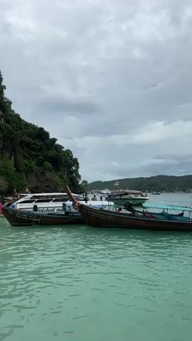 The video shows a body of water with several longtail boats docked near a rocky cliff face covered in lush green vegetation. The water is a vibrant turquoise color. Several people are visible on a sandy beach, some wading in the water near the boats, and others standing on the shore. The sky is overcast with clouds. The boats appear to be for passenger transport, with some having the names of companies or individuals visible on them. The overall scene suggests a tourist destination, possibly an island or a coastal area.