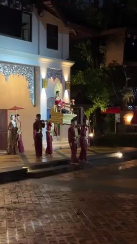 Six men in burgundy and gold traditional attire are carrying a golden palanquin with a person dressed in elaborate white and gold clothing seated on it. Two people in traditional clothing with red umbrellas are standing to the left of the palanquin. The scene takes place at night in front of a white building with ornate gold trim. The ground is paved with bricks. The overall atmosphere appears to be a ceremonial procession.
