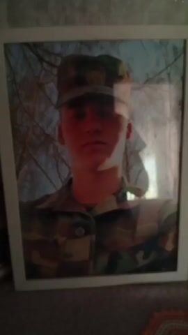 The video shows a framed portrait of a young person in a military uniform. The person is wearing a camouflage uniform and a matching cap. The background of the portrait appears to be an outdoor scene with trees. The lighting in the video is dim, and there are reflections on the glass of the frame. The video is shaky and the focus is not sharp. There is a voice speaking in the background, but it is muffled and unclear.