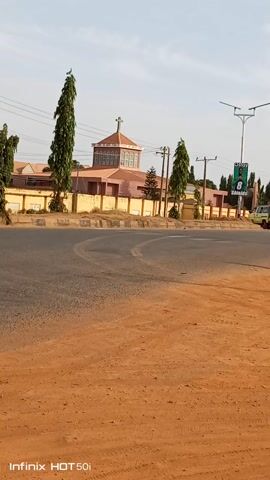 The video shows a street with a church on the left side, viewed from a low angle, and with an orange colored dirt field to the right of the road. The church building is a faded peach color with a cross on top. There are several slender, tall trees lining the property, and a few electrical poles nearby. An advertising banner with a green background is visible further down the road. The video has a watermark in the lower right corner.