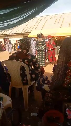 The video captures an outdoor gathering, seemingly a celebration, under a makeshift tent or canopy. Several women are the focal point, adorned in colorfully patterned outfits, and standing amidst a crowd of onlookers. There are a few children in the immediate vicinity. The tent provides partial shade, with buildings visible in the background under bright daylight. A white plastic chair sits prominently in the foreground, and other objects, including beverage bottles, are scattered on the ground, indicative of the social event taking place.