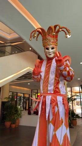 A person dressed in an orange and white harlequin costume with a mask and jester hat makes a peace sign with both hands. The person is standing in a shopping mall. There are plants, escalators, and light fixtures visible in the background.