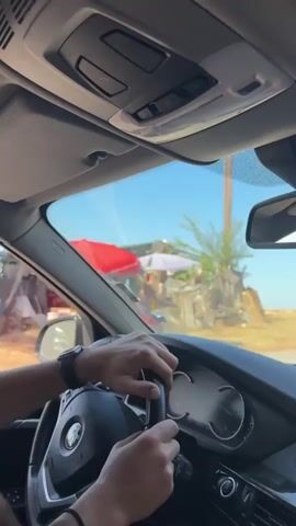 The video shows a first-person perspective from inside a car. A person's hands are visible on the steering wheel and gear shift. The person is wearing a watch on their left wrist. The car appears to be moving along a road, with trees and a body of water visible outside the windows. The sky is clear and blue. The interior of the car is typical of a modern vehicle, with a dashboard and rearview mirror. The scene suggests a leisurely drive through a scenic area, possibly a coastal region.
