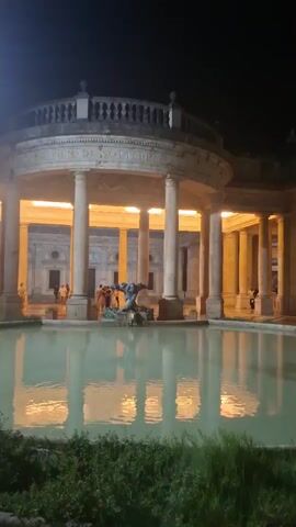 The video shows a nighttime scene of a neoclassical building with a circular colonnade and a domed structure at the top, illuminated by warm yellow lights. In front of the building is a large, still pool of water reflecting the lights and the structure. In the center of the pool, a dark statue of a human figure can be seen. Several people are gathered in the courtyard area behind the pool, some appearing to be interacting. The foreground is partially obscured by dark green foliage. The lighting suggests it is evening or night.