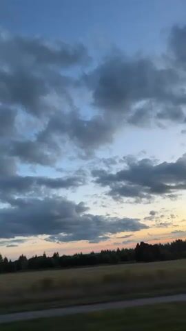The video shows a time lapse of clouds moving across a blue sky. The clouds are dark and fluffy, and they are illuminated by the setting sun, which casts a warm glow on the horizon. Below the sky, there is a line of trees and a field of grass. The overall scene is peaceful and serene.