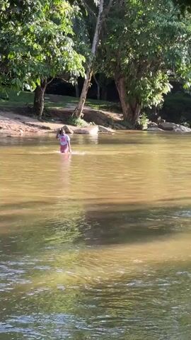 A group of children and an adult are wading through a shallow, murky river surrounded by lush green trees and rocky banks. Sunlight filters through the leaves, casting dappled reflections on the water's surface. The children appear to be playing and splashing, with one child holding a pink inflatable ring. The adult is carrying a young child. The overall atmosphere is one of relaxed outdoor recreation.