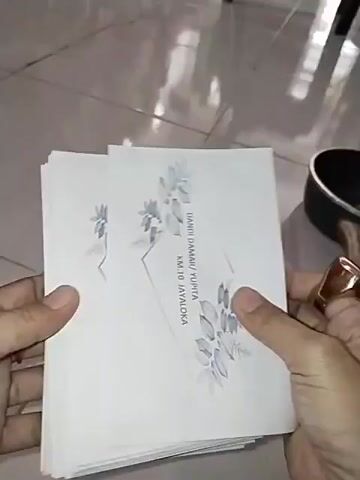 A close-up shot shows a person's hands holding a stack of white envelopes with a grey floral design. The envelopes have text printed on them, including names and an address. The person is flipping through the envelopes, revealing the text on each one. The background is blurry and appears to be a tiled floor.