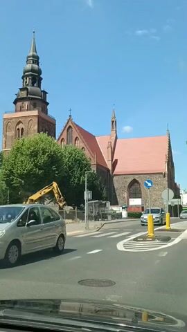 A video captures a sunny day in Gryfino, Poland, with a prominent brick church as the central focus. The church features a tall, dark spire, a red-tiled roof, and arched windows. Adjacent to the church, a construction site is visible, complete with a yellow excavator and a pile of dirt.

The scene unfolds on a street in front of the church, where traffic moves along a paved road marked with white lines and a circular intersection. A silver car navigates the intersection, followed by another car. Yellow bollards and a blue directional sign indicate traffic flow.

The weather appears to be clear and bright, suggesting it's daytime. The presence of trees with lush green foliage indicates a warm season, likely spring or summer. The overall atmosphere is one of urban activity with a backdrop of historical architecture. No legible text is clearly visible in the video.
