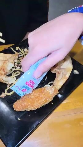 This media file captures a close-up of a person's hand squeezing a packet of "Bangkok Sauce" onto a fried food item, likely a fish stick or chicken tender, which rests on a black plate. The plate also contains a portion of noodles and some fried dumplings. The food is placed on a wooden table, and in the background, a hint of a person wearing a black shirt is visible.

The activity is the act of seasoning food with a sauce packet, suggesting a meal is being prepared to be eaten. The lighting appears to be indoor, possibly from overhead lights, and no information about the time of day or weather is available. The setting seems to be a casual dining environment, possibly a restaurant or a home. The hand holding the sauce packet is the primary focus, with the texture of the fried food and the white and pink packaging of the sauce clearly visible. The visible text on the packet reads "Bangkok Sauce" in stylized lettering. The overall impression is a simple, everyday action of enjoying a meal.