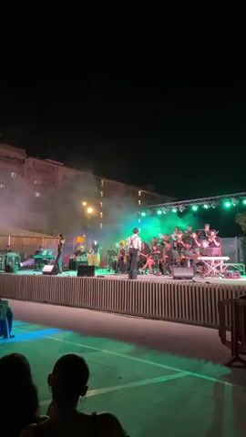 The video shows a live outdoor concert at night. A band is performing on a brightly lit stage, with a singer in the foreground. The audience is seated in front of the stage, with some people closer to the stage and others further back. The backdrop of the concert is a building with lit windows, and trees are visible in the foreground. There is smoke or fog on stage, illuminated by the stage lights. The lighting is predominantly green and white.