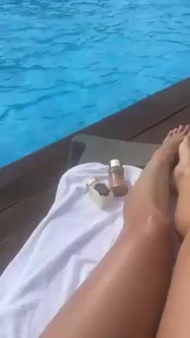The video shows a person relaxing by a pool. The camera is positioned looking down at the person's legs resting on a white towel on a gray lounge chair. There are two small bottles on the towel next to the person's legs. The pool is a bright blue color and people are swimming in it. In the background are forested hills and a partly cloudy sky. The pool has a wooden deck surrounding it. There are voices and sounds of people in the background.