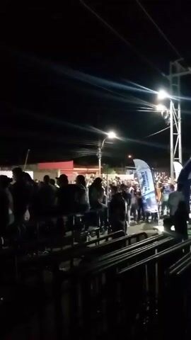 The video shows a concert at night. The camera is initially shaky and focuses on a crowd of people in front of a fence or railing, then pans to the right, revealing a brightly lit stage. A band is performing on the stage. The crowd is silhouetted in the darkness. The setting appears to be outdoors, with streetlights visible in the background.