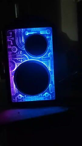 The video displays a close-up, low-light shot of a black, rectangular object with two circular openings, resembling a speaker or an electronic device. The object is illuminated from the side and from within by LED lights that cycle through colors, starting with purple and transitioning to various shades of blue. The surface of the object features an intricate, etched design of interconnected lines, circles, and gear-like shapes, giving it a futuristic or steampunk aesthetic. The lighting casts a glow on the surface and a diffuse light onto the dark, indistinct background, which appears to be a surface such as a table. The camera angle is slightly from above and to the side, focusing on the illuminated design and the glowing openings. There are no people or discernible actions other than the color-changing illumination of the object.