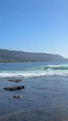 The scene shows a surfer riding a wave in the ocean on a bright, sunny day. The water is a clear blue-green, and the waves are moderately sized with white foam. The surfer is wearing a dark wetsuit and is actively riding the wave. In the foreground, there are several dark rocks partially submerged in the water, adding depth to the scene. The background features a lush green mountain range that stretches along the coastline, with some buildings visible at the base of the mountains. The sky is a clear blue with a few puffy white clouds. The camera is static, providing a fixed view of the surfing activity and the surrounding landscape.
