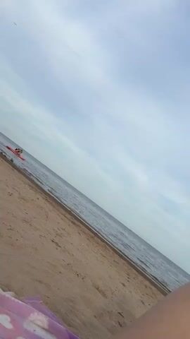 The video shows a wide shot of a beach and ocean under a cloudy sky. In the water, a person is on an inflatable red raft, appearing to be paddling. Another person is also visible on the raft. The water is calm with small waves lapping the shore. The sand on the beach is light brown and appears dry. The camera is tilted, giving the scene a dynamic angle.