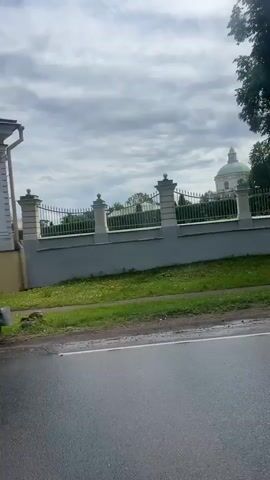 The video shows a drive-by view of a white stone fence with decorative posts and an ornate metal gate. A large white building with a green dome is visible behind the fence. Two people are walking on a wet asphalt road in front of the fence. One person is wearing a blue shirt and gray pants, and the other is wearing a light-colored jacket and pants, carrying shopping bags. The sky is overcast. A white car is parked on the right side of the road. The video captures the scene in a series of short clips, suggesting movement through the area.