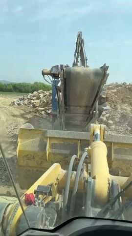 The video shows the perspective from inside the cab of a large yellow loader at a construction site. The loader is positioned in front of a rock crusher, which is being operated by a worker in a red vest. The camera pans around, showing the dusty environment and the other heavy equipment in the area, including an excavator. Piles of rocks and dirt are visible in the background, and the loader appears to be moving into position to begin work.