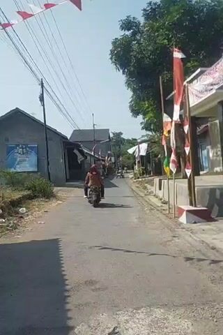 A person is riding a motorcycle down a narrow, paved road. The road is lined with simple houses, some of which have Indonesian flags displayed. Trees and power lines are visible in the background. The sky is clear and blue. Another motorcycle is visible further down the road. The scene appears to be a rural or suburban neighborhood.