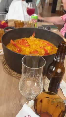 The video shows a close-up shot of a large, black pot of lobster paella on a wooden table in a restaurant setting. The paella contains bright red lobster pieces and yellow rice in a rich broth. A clear glass, a bottle of beer, and woven placemats are also visible on the table. In the background, other diners are partially visible, along with glasses of drinks, sunglasses, and other items. The camera focuses on the paella, highlighting its vibrant colors and the presentation of the lobster. The overall scene suggests a meal being shared among diners in a casual, restaurant environment.