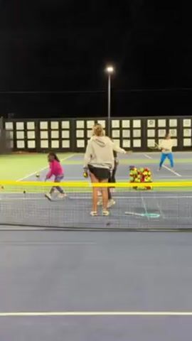 The video shows several individuals on a tennis court at night.  The court is blue with green sidelines. There's a standard tennis net separating the halves. A light pole illuminates the court. Behind the court, there's a black fence with white rectangles on it. One person, wearing a pink top and patterned leggings, is actively hitting tennis balls. Another person is wearing blue pants and a white top. Three other people, dressed in more casual clothing, stand near a wire basket filled with tennis balls, presumably coaching. They offer instructions and feedback after each shot. One coach is wearing a grey sweatshirt, one is wearing a white sweatshirt, and the third is partially obscured. Tennis rackets are visible near the basket.