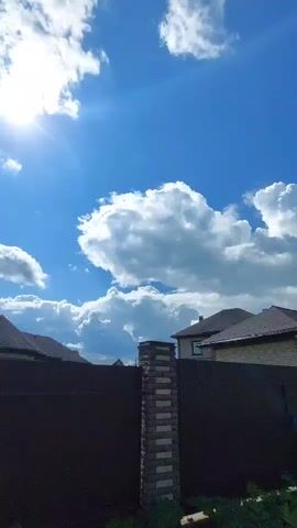 This is a vertical video of a suburban neighborhood on a sunny day. The sky is a vibrant blue with large, fluffy white clouds. The sun is visible in the upper left corner, casting rays of light. In the foreground, there is a dark brown wooden fence with a brick pillar on the left. Beyond the fence, several houses are visible, some appearing to be under construction with exposed framing. Green foliage and grass are present in front of the fence. The camera pans slightly to the right, revealing more houses and the fence line.