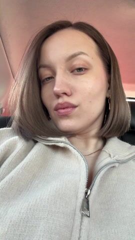 The video shows a woman from the shoulders up, sitting in the backseat of a car. She has pale skin, blue eyes, and short brown hair. She is wearing a beige zip-up sweater and gold earrings. She looks directly at the camera with a slightly pouting expression. She touches her hair and earring. The car's interior is visible, with a black leather seat and a red-tinged roof.