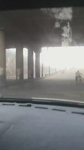 A vehicle is traveling on a road under a bridge. The road is lined with concrete barriers and streetlights. The sky is bright and hazy. There are other vehicles on the road, including cars and a motorcycle. The view is from inside the vehicle, looking out the windshield. The dashboard of the vehicle is visible in the foreground. The scene appears to be in an urban or semi-urban area.