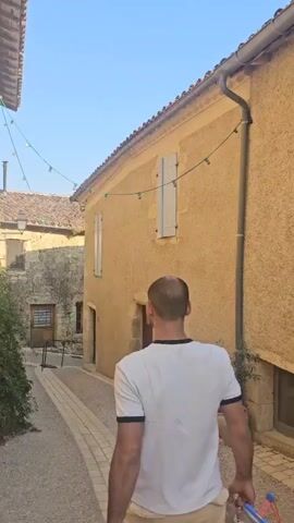 A man wearing a white t-shirt and light-colored shorts walks away from the camera down a cobblestone path. A woman in a light-colored top and dark shorts walks beside him. They are walking alongside a textured yellow building with blue shutters and a drainpipe. String lights hang across the building. There is a small potted plant next to the building. Further down the path, there is a blue door, more potted plants, and a second window with a hanging object made of metal. The path continues into the distance, lined by similar buildings.