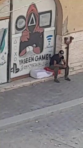 The video shows a man sitting on a large white bag on a sidewalk. He is wearing a dark t-shirt, dark pants, a baseball cap, and dark shoes. He is looking down and appears to be resting. Behind him, there is a wall with graffiti on it, including a drawing of a character from the game Squid Game, the PlayStation logo, and text in Arabic. There is also a metal pole with a curved top to the right of the man. The ground in front of him is paved with gray stones. The lighting suggests it is daytime.