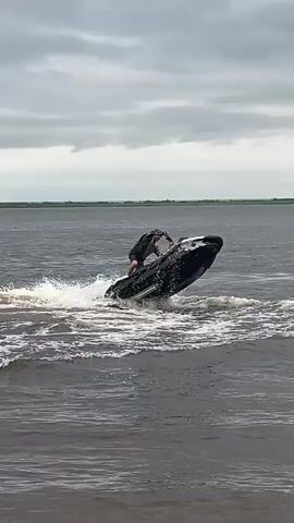 A person rides a jet ski in a body of water. The jet ski is black and red, and the person is wearing a dark jacket and shorts. The water is choppy and there is a lot of spray. The sky is overcast. The person appears to be making a sharp turn.