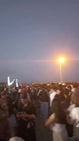 The video shows a large crowd of people at an outdoor event under a dusk sky. There is a stage in the distance with bright lights and fog. People are standing close together, facing the stage. Some are looking at their phones. A few trees are visible in the background.