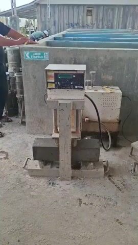 A concrete testing machine is shown. The machine has a digital display and a hydraulic press. Two concrete beams are placed on the machine's base. A person's hands are visible on the left side of the frame, resting on a concrete wall. In the background, there are several large, blue, rectangular containers. The environment appears to be an outdoor industrial or construction site, with concrete surfaces and a weathered building in the background. The machine is powered on, with lights and numbers visible on the display. The video shows the machine in operation, but no specific test is being performed.