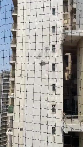 A daytime video shows the exterior of a multi-story apartment building in Noida, India, viewed through a black safety net in the foreground. The camera pans slowly from left to right, revealing the building's facade. The building has a light-colored exterior, possibly off-white or cream, with some visible wear and tear, including discolored patches on the walls. Numerous windows and balconies with metal railings are visible. Clothes are seen drying on a rack on one of the balconies. Air conditioning units are also mounted on the building's exterior. The sky is clear, indicating good weather. No people are clearly discernible.