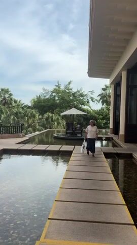 A woman is walking on a stone walkway that spans across a shallow pool of water. She is wearing a white shirt, a dark skirt, and a headscarf. She is carrying a bag in her left hand. The walkway leads to a small circular island in the middle of the pool, where there is a table and chairs under an umbrella. The background shows lush green trees, palm trees, and a cloudy sky. A modern building with a white roof and pillars is visible on the right side of the frame. The water in the pool is clear, and pebbles are visible at the bottom. The overall scene appears to be an outdoor courtyard or garden area.
