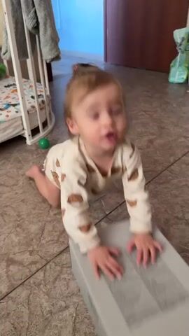 A baby is on its knees on a tiled floor, wearing a beige onesie with brown teddy bear prints. The baby has its hands on a gray box in front of it, and its hair is pulled up into a small ponytail. The baby looks at the box and then claps its hands together. In the background, there is a crib, a door, and some other objects.