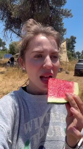 A young woman with brown hair tied up in a bun and wearing a grey sweatshirt is eating a slice of watermelon. She is holding the watermelon in her right hand and taking a bite. Her eyes are closed as she chews. The background shows a dry, grassy field with trees and a tent. There is also a car parked in the distance. The lighting suggests it is a sunny day.