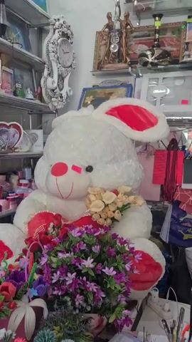 A white stuffed rabbit toy sits in a shop cluttered with various items, including framed pictures, a large ornamental clock, and colorful decorative flowers. The rabbit is holding a small bouquet of beige flowers and has a larger bouquet of purple and pink flowers in front of it. The toy has red accents on its ears and feet. The background appears to be a shelf filled with more items, and there are shopping bags and other merchandise visible.
