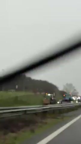 The video shows a line of tractors driving along the shoulder of a highway. The tractors are various colors, including green, blue, and gray. Many of the tractors have orange warning lights flashing. Some tractors have signs that say "STOP LANDGRAB." The weather appears to be overcast and gray. The video is taken from inside a vehicle moving alongside the tractors. There is a line of trees on the right side of the highway.