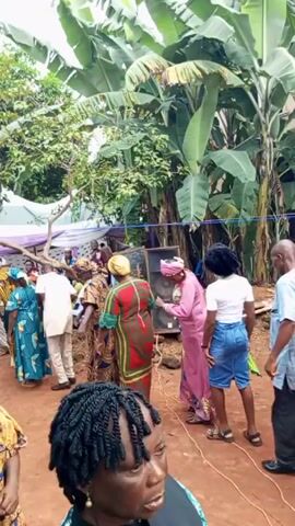 The video captures a lively outdoor gathering with a mix of people, mostly adults, dressed in colorful traditional African clothing. They appear to be part of a celebration or event, moving about in a social manner. The background shows lush greenery, banana trees, and a partially visible white tent with purple trim, suggesting a festive outdoor setup. The camera moves to follow the crowd, offering glimpses of faces and outfits, creating a sense of participation in the event.