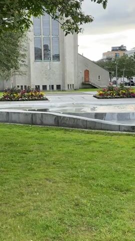 The video shows a gray, two-story building with large windows and a small orange door with a staircase leading to it. In front of the building is a fountain with two seagulls. To the right of the fountain is a large stone statue of a walrus. A person wearing a light blue shirt and pants is standing next to the walrus statue. Behind the fountain, there is a flower bed with red, yellow, and pink flowers. In the background, there are trees, buildings, and parked cars. The sky is overcast.