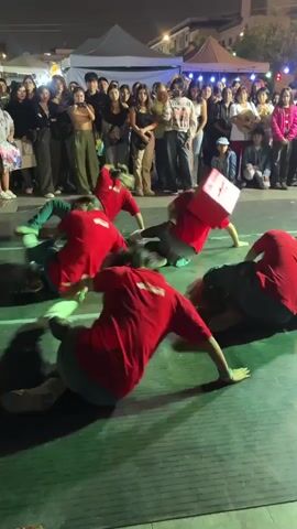 A group of young women in red shirts and denim shorts are performing a synchronized dance routine on a paved outdoor surface at night. They are seated on the ground in various positions, some with legs extended, others with knees bent. Their hair is long and dark, and it is flying around their faces as they move. The dancers are facing a crowd of spectators who are standing behind a barrier. The crowd is diverse in age and gender, and they are watching the performance with interest. Some people in the crowd are holding up their phones to record the performance. In the background, there are tents and streetlights, suggesting that the performance is taking place at an outdoor event or festival. The lighting is dim, and there are some colored lights illuminating the dance floor.