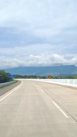 This is a first-person perspective video of driving on an empty, paved highway. The road curves to the left and stretches into the distance, disappearing behind a hill. On the left side of the road, there are trees and a blue sign. On the right side of the road, there are yellow curve warning signs and a white barrier. In the background, there are mountains and a cloudy sky. The weather appears to be overcast.