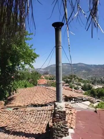 The video shows a close-up view of a metal chimney on a tiled roof. In the background, there is a village nestled in a valley with rolling hills and a clear blue sky. The chimney appears to be old and weathered, with wires attached to it, likely for support. The roof tiles are also old and some appear to be missing or broken. The overall scene suggests a rural and somewhat rustic setting. The video pans slightly, offering a wider view of the village and the surrounding landscape. There are no people or animals visible in the video. The sunlight is bright, indicating it is daytime.