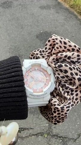 A close-up shot captures a person's wrist, featuring a white Casio Baby-G watch with a rose gold/pink face. The watch dial shows "LON" for London and "MON 9:56", indicating the time and day. Various inscriptions like "WORLD TIME", "SHOCK RESIST", "PROTECTION", and "CASIO WR 10BAR" are visible on the watch. The wearer's black ribbed knitted sleeve is partially visible on the left, while a leopard print scrunchie, light brown with black spots, is wrapped around the wrist on the right side of the watch. The background shows a blurry paved surface, likely a road or path, with a hint of green vegetation in the lower left corner, suggesting an outdoor setting. The camera holds steady on the subjects throughout the short video.
