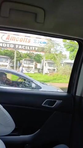 The video shows a view from inside a moving car, looking out the rear passenger side window. A silver car can be seen driving next to the camera car. The driver of the silver car is visible, appearing to be a Black male talking on a mobile phone. Beyond the silver car, there are various roadside elements like an Exxon sign, parked trucks, and trees. As the car moves, the scenery changes to show fences, buildings, and more trees.  The interior of the camera car is visible, featuring dark colored upholstery, a door handle, and a headrest.