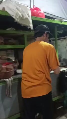 A man in an orange t-shirt and black cap is shown from the back, reaching out to the right. He is standing in front of a food stall at night. Behind him, a white bucket sits on a green stool. To the right, an older man in a white shirt and a black cap, wearing a green sarong, is holding a small white object and a plastic bag. A woman in a black hijab is partially visible in the foreground to the right, looking towards the older man. The background shows a street scene at night with vehicles and other stalls. Decorative flags are strung above the stall.