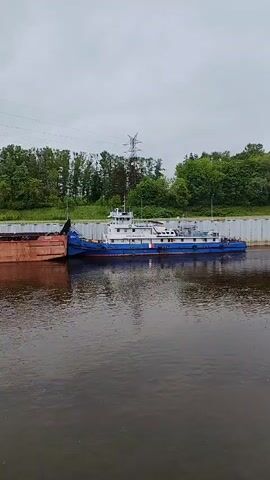 A static shot shows a blue and white tugboat pushing a rusty red barge on a body of water. The tugboat is long and rectangular with a small superstructure. The barge is shorter and filled with a dark material. The water is dark and reflects the sky. The canal is lined with concrete walls. In the background, there is a line of trees and a gray sky.  There are also power lines and street lights visible.