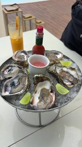 The video shows a plate of oysters on ice, a glass of orange juice, hot sauce, and pink sauce on a white table. Behind the table is a blue swimming pool with a wooden deck, leading to a beach with umbrellas and the ocean in the background. A white building is visible on the right. There is also a large white planter and a stack of gold lanterns near the pool.