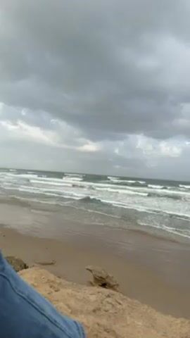 The scene displays a sandy beach under an overcast sky. Waves break along the shoreline. In the foreground, a person wearing jeans is visible, seemingly sitting on a rocky or sandy elevation.